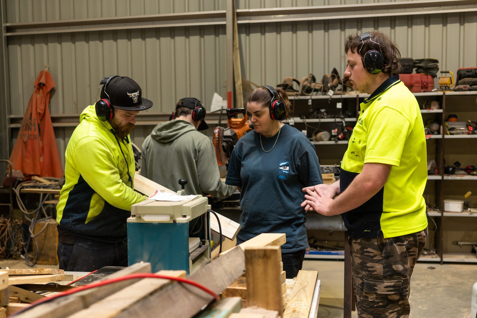 Group of people wearing safety earmuffs working together in a woodworking workshop, surrounded by timber, tools, and machinery.