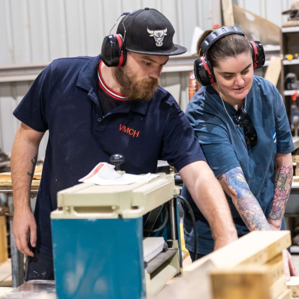 Two people wearing protective earmuffs working together in a woodworking workshop, operating machinery and handling timber.