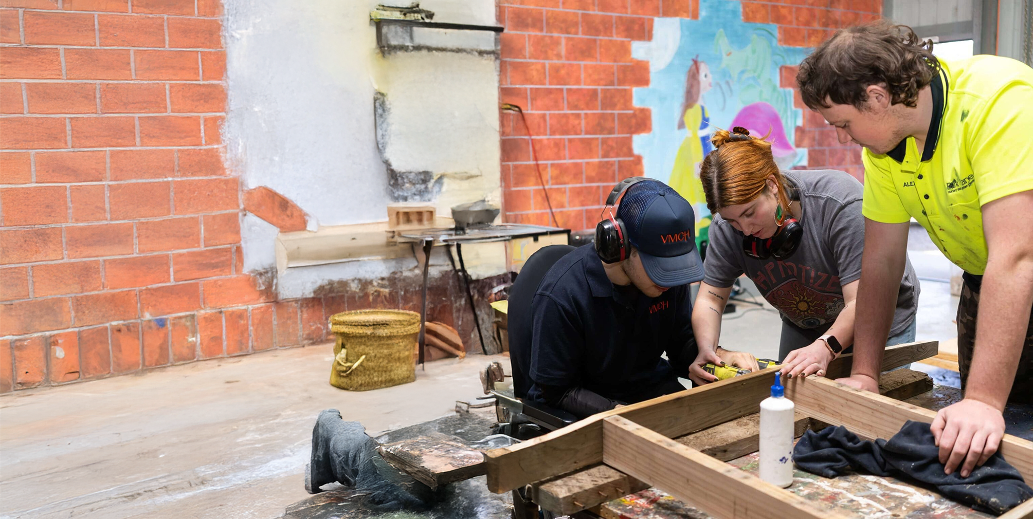 Three people working together on a woodworking project in a workshop, with tools and timber on the table, as part of a VMCH disability services program.