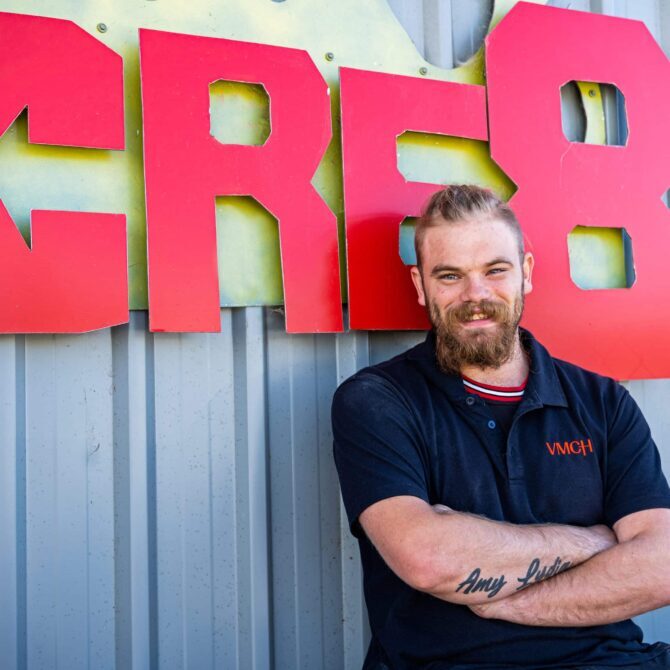Man wearing a VMCH polo shirt smiling with arms crossed in front of a large red “CRE8” sign on a corrugated wall.